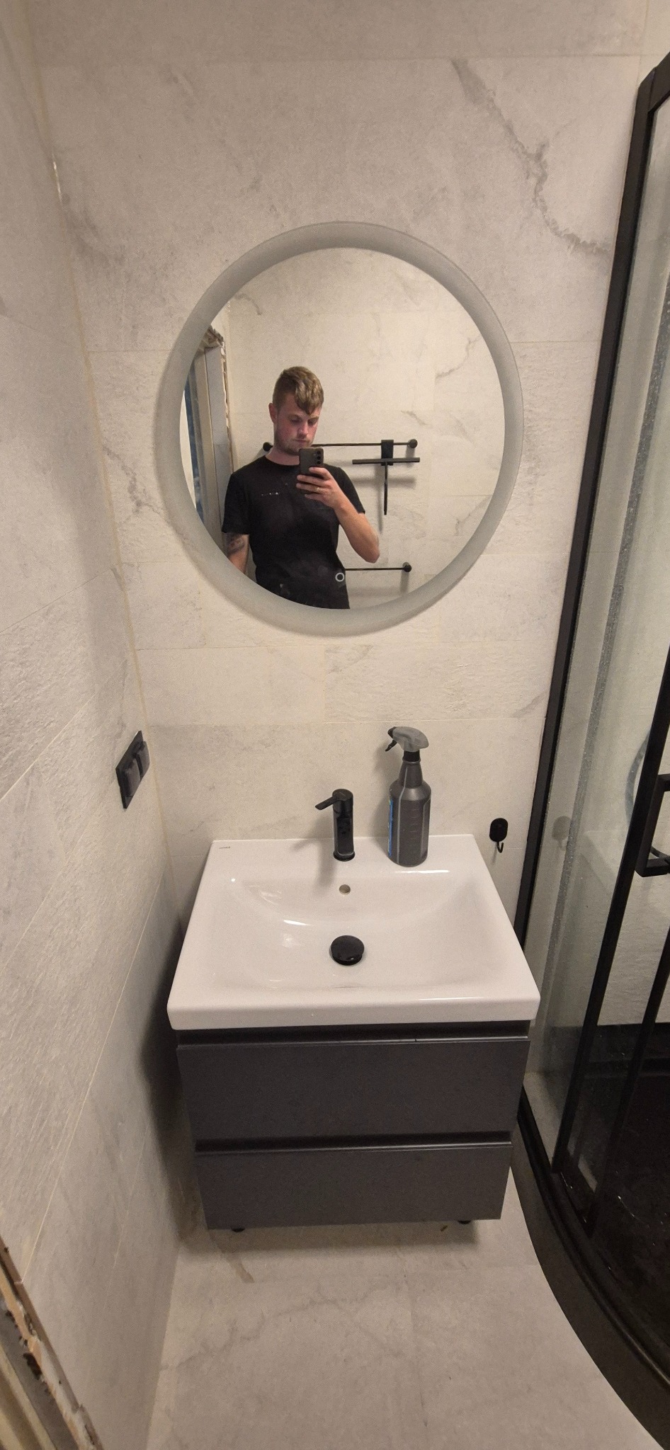 Nowoczesna łazienka z okrągłym lustrem, białą umywalką z szarą szafką i kabiną prysznicową z czarnymi ramami. Widoczne płytki imitujące marmur.