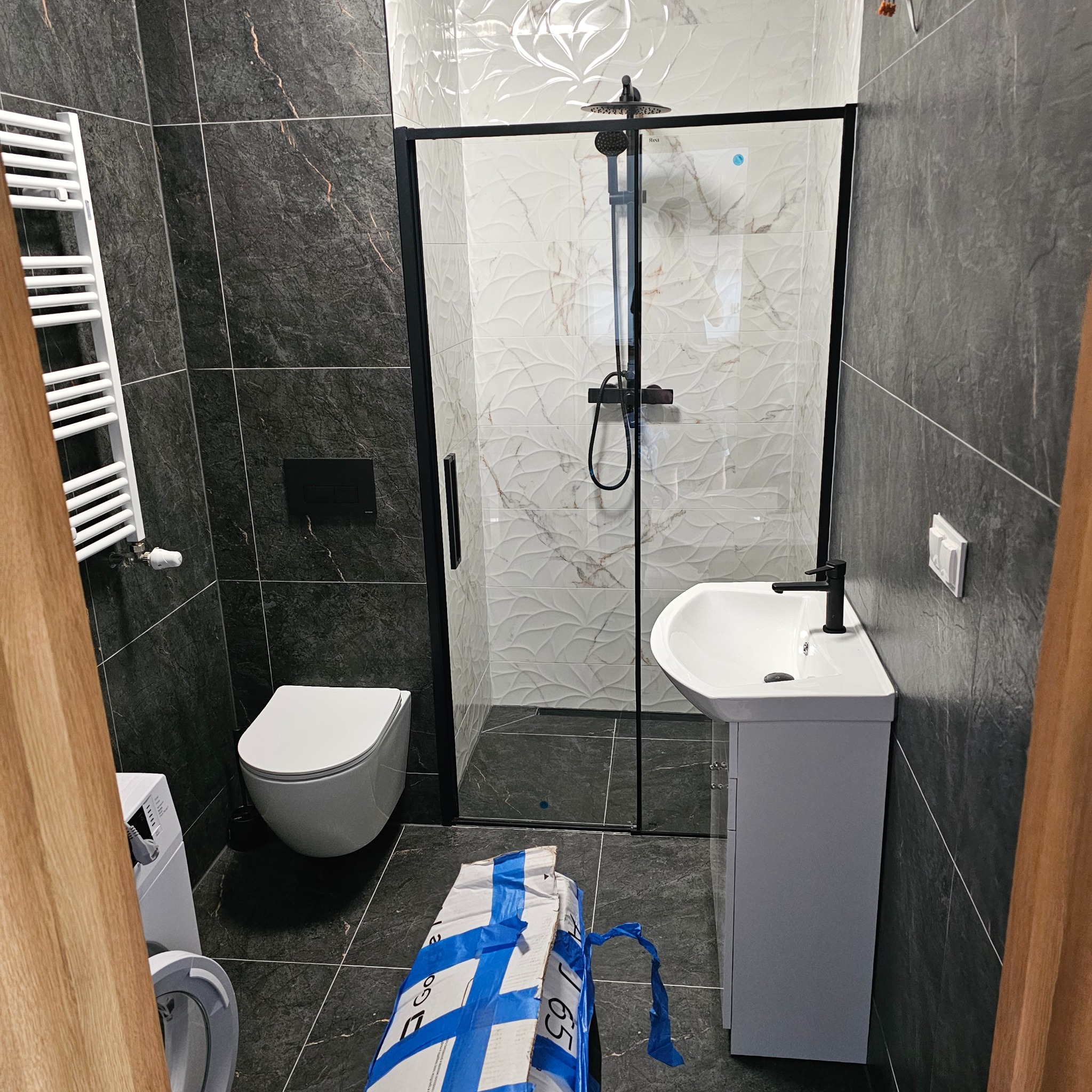 Nowoczesna łazienka po remoncie: kabina prysznicowa z czarną ramą, umywalka z szafką, toaleta i grzejnik drabinkowy na tle ciemnych płytek. Widoczny fragment pralki.