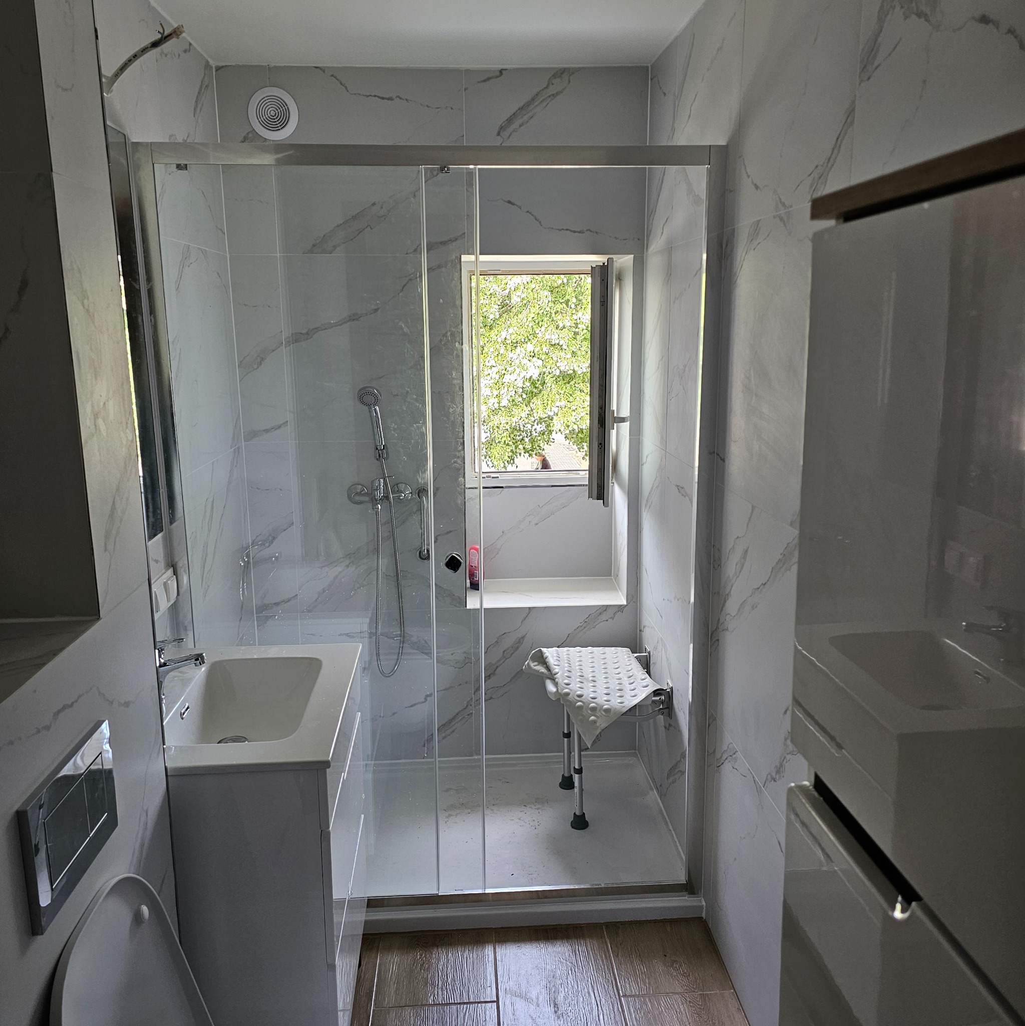 Nowoczesna łazienka z kabiną prysznicową, marmurowe płytki na ścianach, drewnopodobna podłoga, okno z widokiem na zieleń. Minimalistyczny design, jasne kolory.