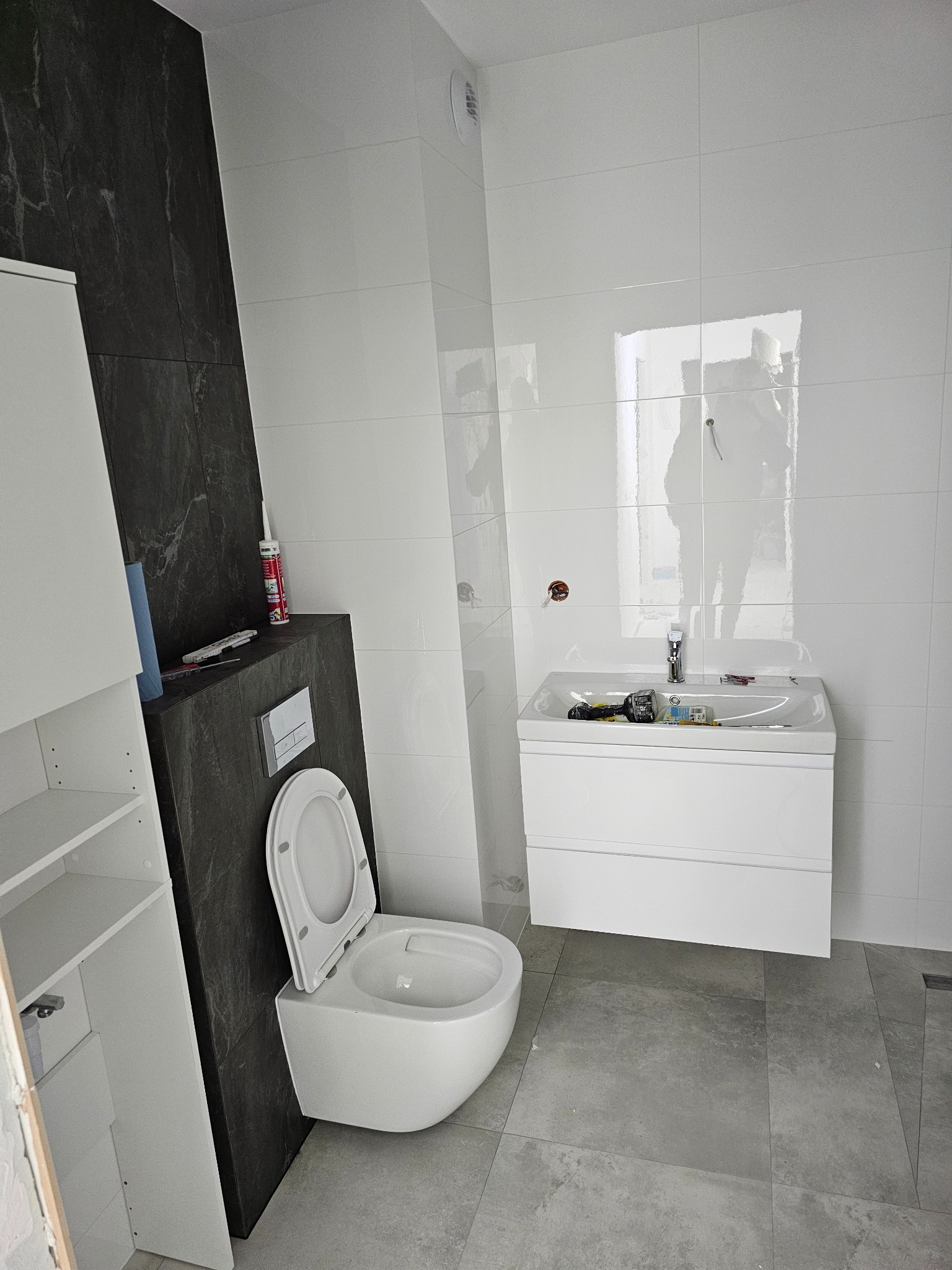 Nowoczesna łazienka w trakcie wykańczania: białe płytki, szara podłoga, podwieszana toaleta z otwartą klapą i biała szafka z umywalką. Widoczne narzędzia.