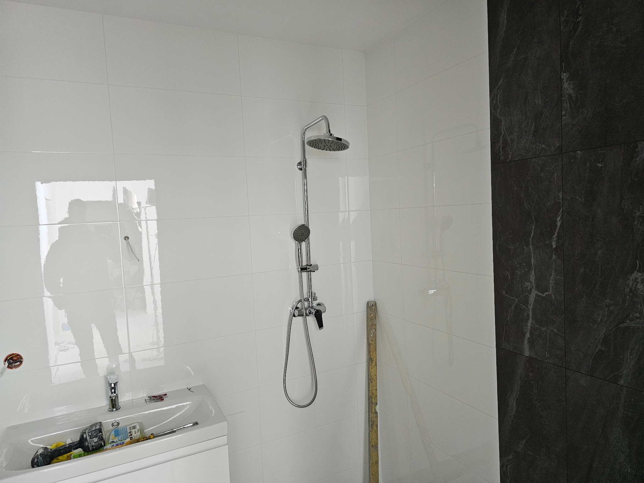 Wykończona łazienka z białą glazurą i czarną ścianą imitującą marmur. Widoczna kabina prysznicowa, umywalka z narzędziami glazurniczymi. Poziomica oparta o ścianę.