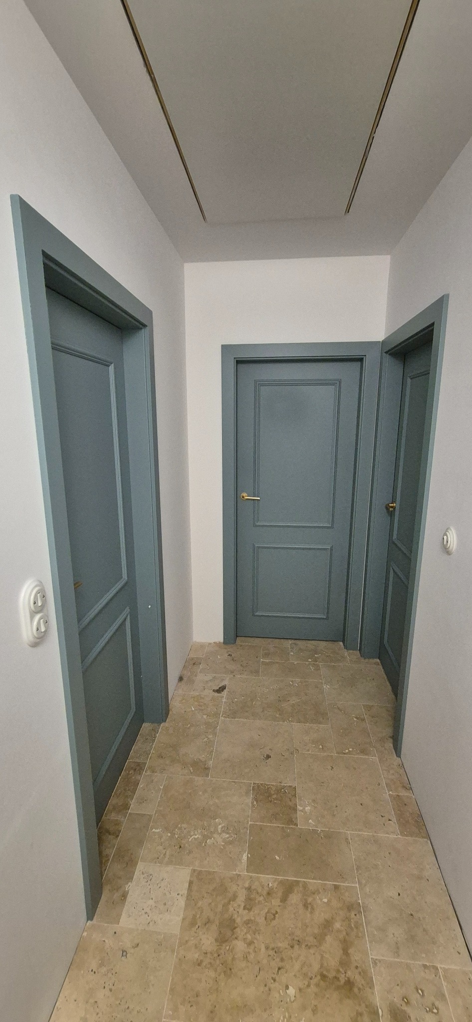 Korytarz z trzema szarymi drzwiami z ozdobnymi ramami i beżową podłogą z płytek. Widoczny włącznik światła i listwy na suficie.