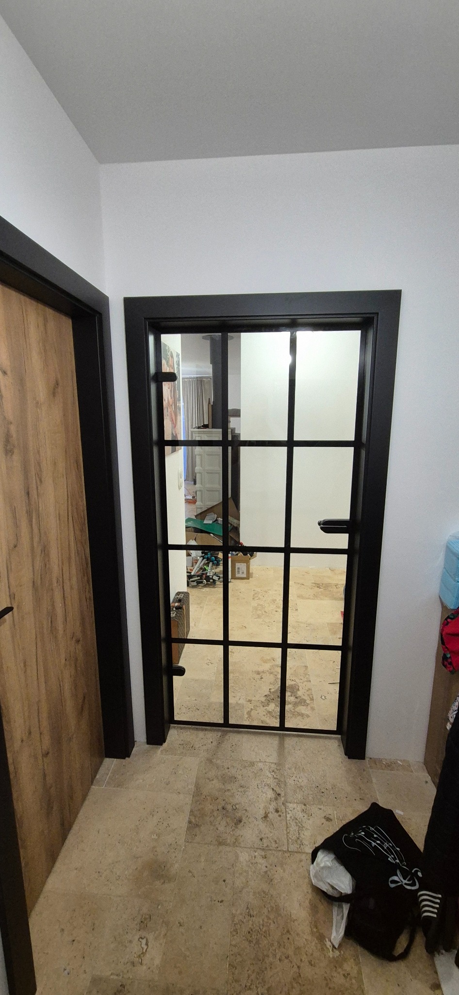 Nowoczesne drzwi wewnętrzne z czarną ramą i szprosami, widok na wnętrze z beżową podłogą. Kontrastujące wykończenie z drewnianymi drzwiami po lewej.