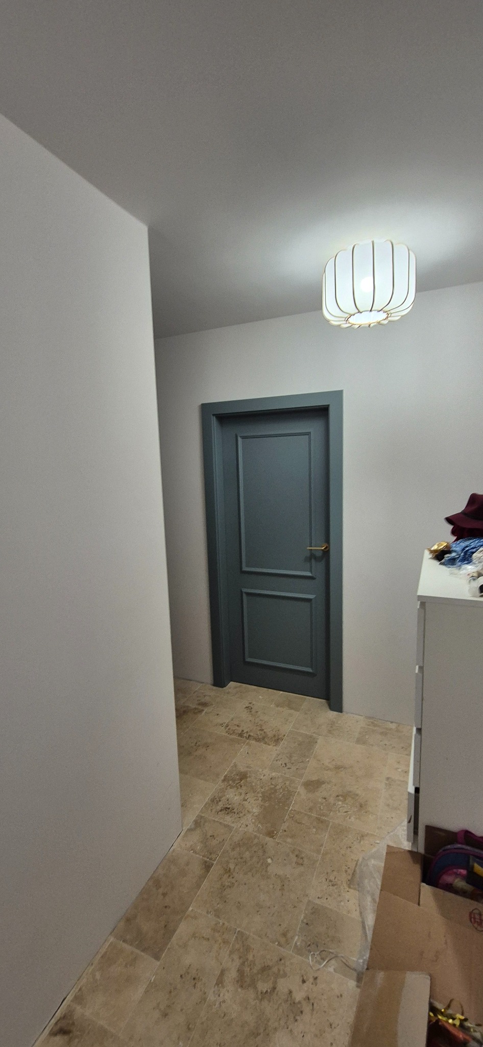 Szaro-niebieskie drzwi z panelami w jasnym korytarzu z beżową podłogą z płytek. Nad drzwiami widoczna lampa sufitowa.