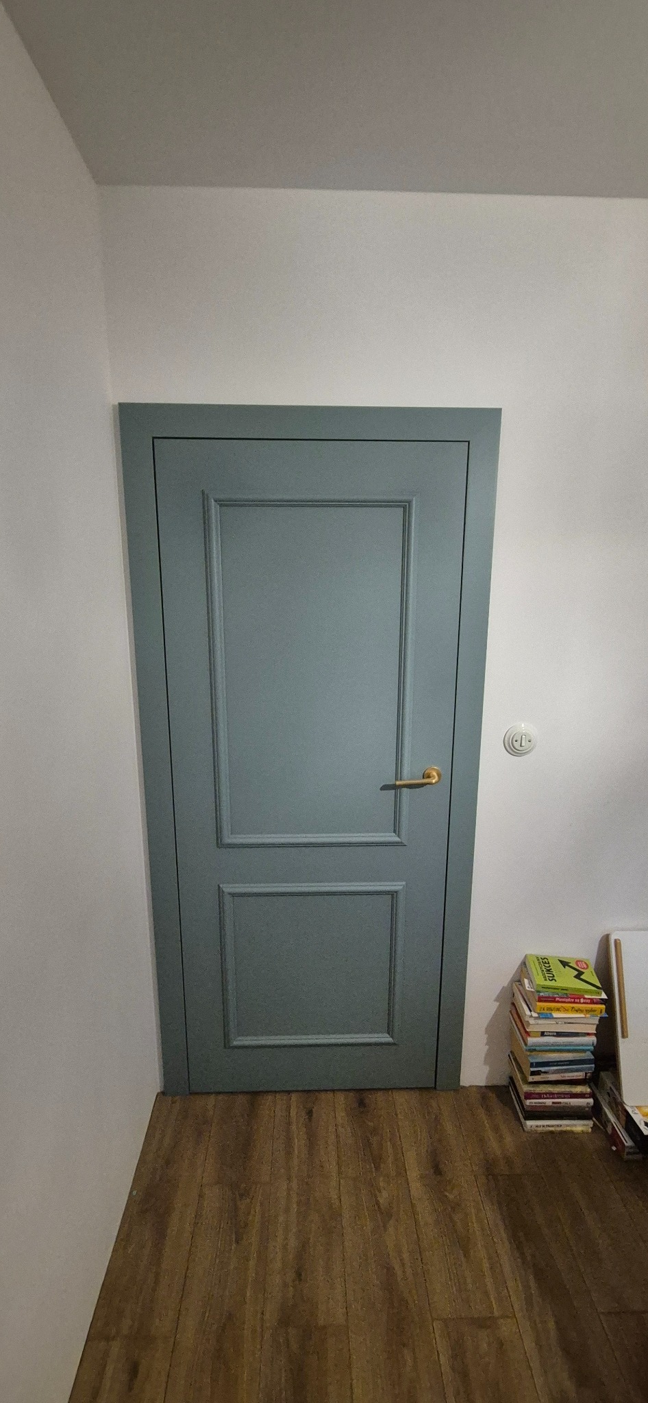 Szara, panelowa, zamknięta  drzwi z złotą klamką, na drewnianej podłodze. Obok ściany stos książek. Widok na wykończone wnętrze.