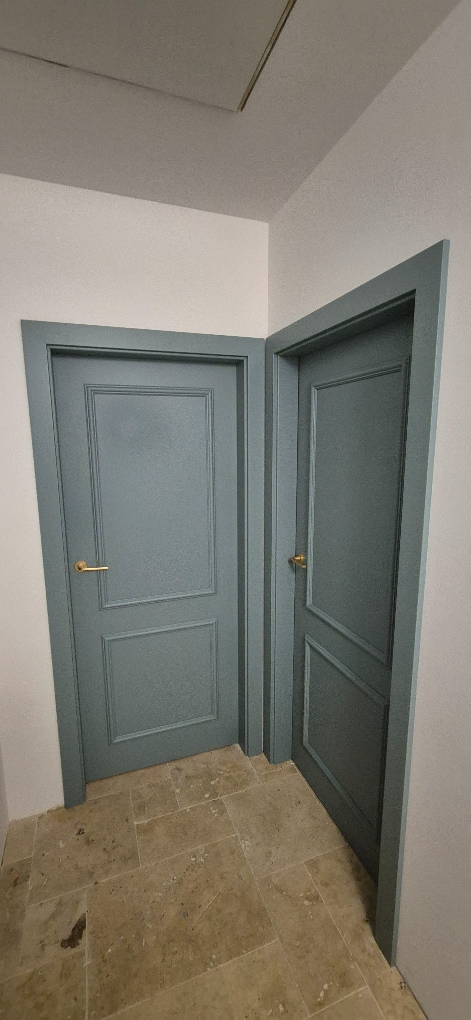 Dwa odcienie szarości: Klasyczne drzwi wewnętrzne z panelami i złotymi klamkami, ustawione pod kątem w jasnym korytarzu z kamienną podłogą.