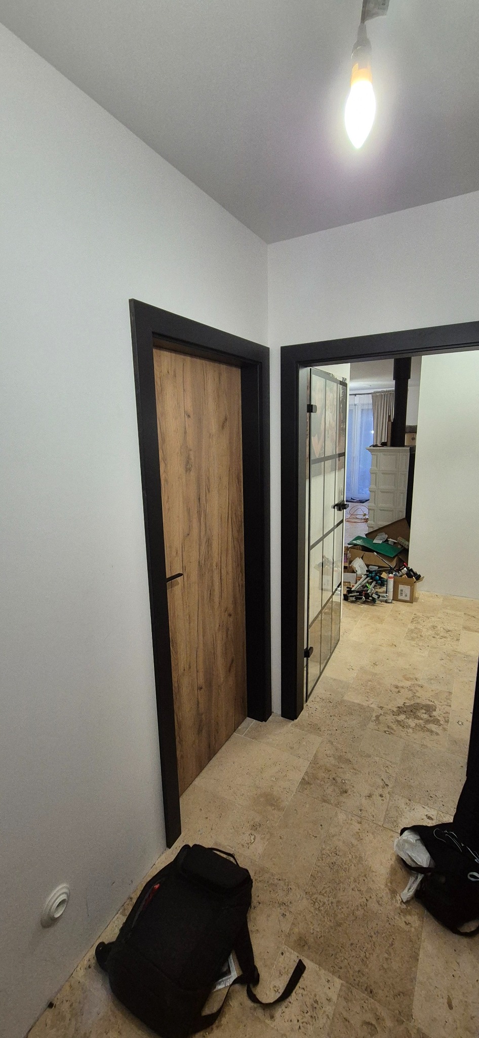 Dwa odnowione wejścia z czarnymi ościeżnicami: jedno z drewnianymi drzwiami, drugie przeszklone. Na podłodze beżowe płytki, w tle widoczne narzędzia.