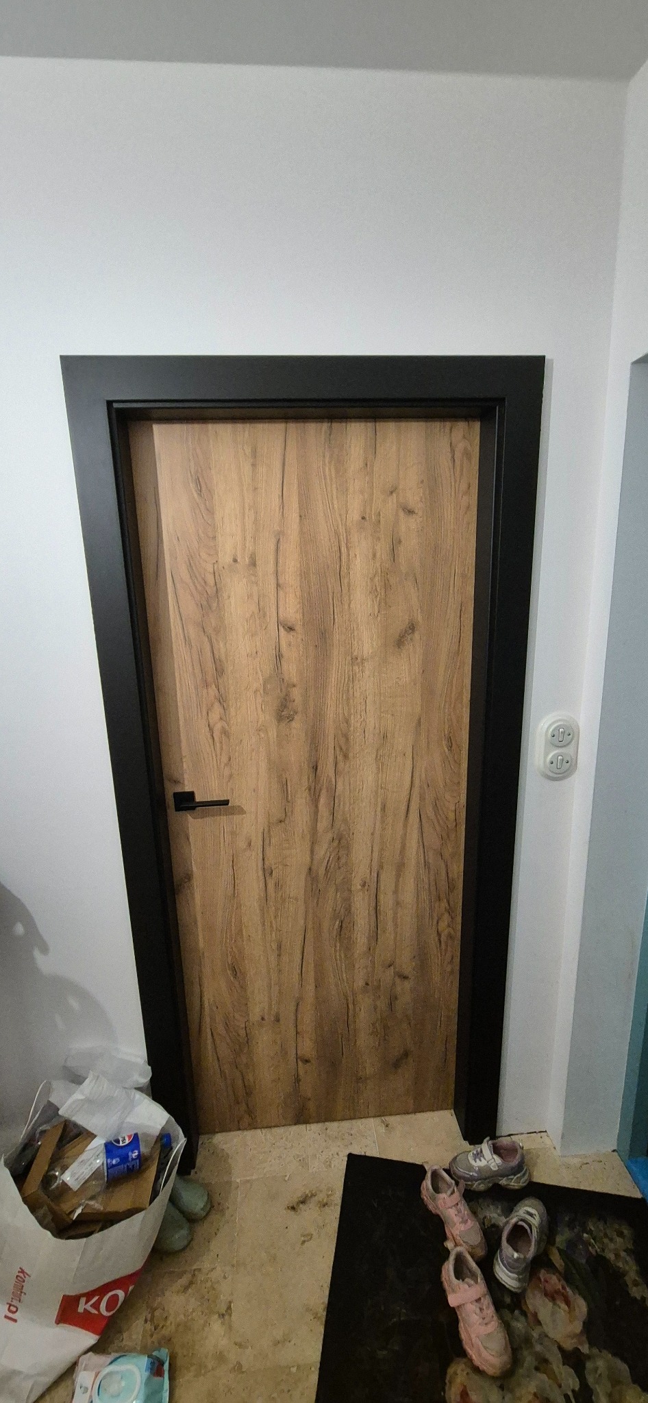 Zamknięte drzwi wewnętrzne z drewnianą okleiną i czarną ościeżnicą, widoczne na tle białej ściany z włącznikiem. Na podłodze torba z odpadami i dziecięce buty.