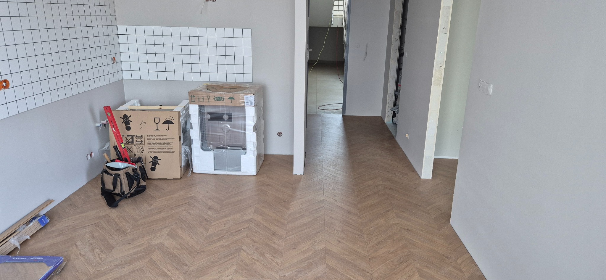 Ukończona podłoga z parkietu ułożonego w jodełkę w jasnym wnętrzu. Widoczne płytki na ścianie, narzędzia i zapakowane urządzenia, sugerujące trwający remont.