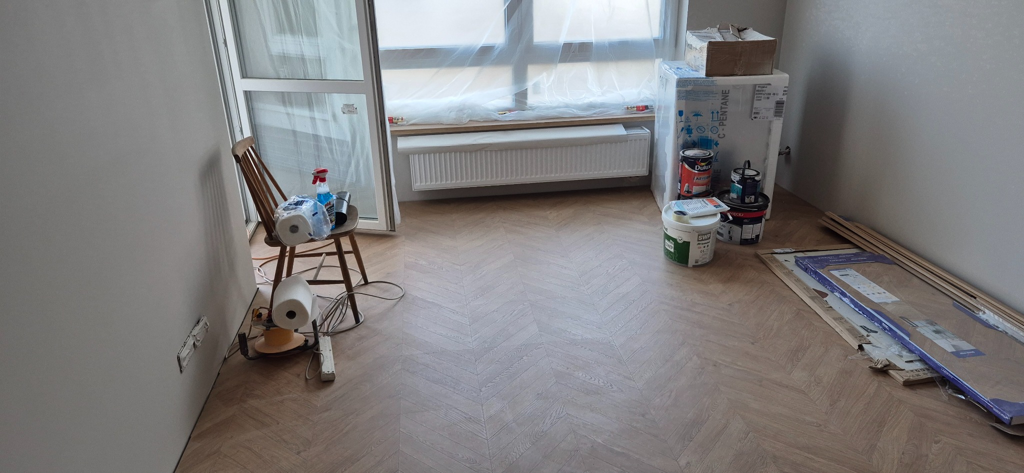 Jasne panele ułożone w jodełkę w pokoju podczas prac wykończeniowych. Widoczne farby, oklejone okno, listwy przypodłogowe i krzesło z akcesoriami malarskimi.
