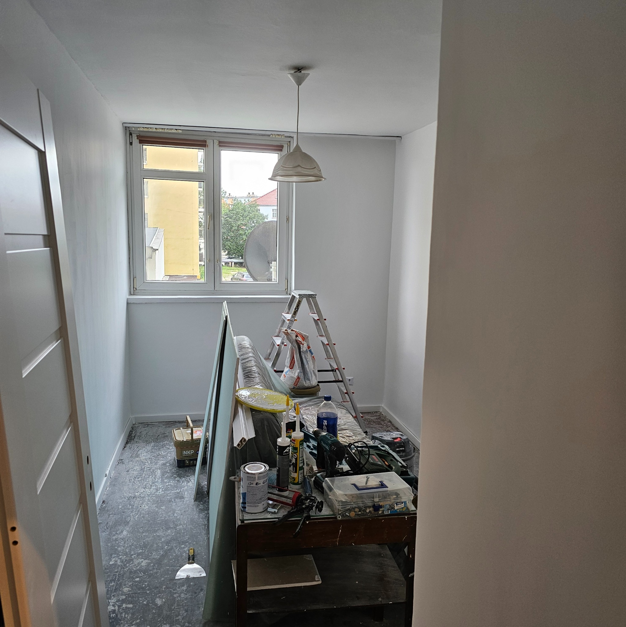 Wnętrze w trakcie odświeżania: białe ściany, okno z widokiem na budynek, drabina, płyty gipsowe i narzędzia na stole. Widoczny etap prac wykończeniowych.