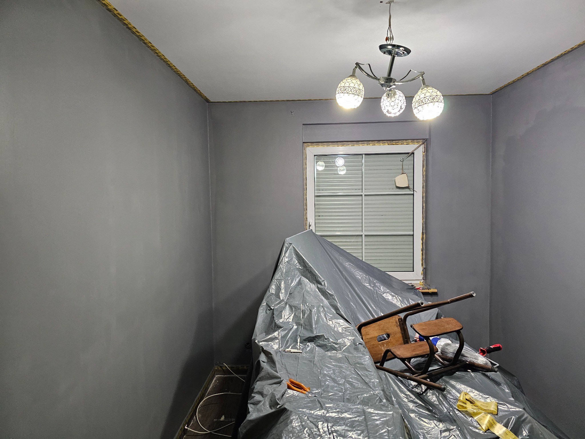 Pokój po malowaniu na szaro, zabezpieczony folią i krzesłem. Widoczna lampa sufitowa i okno. Świeżo pomalowane ściany z żółtą listwą pod sufitem.