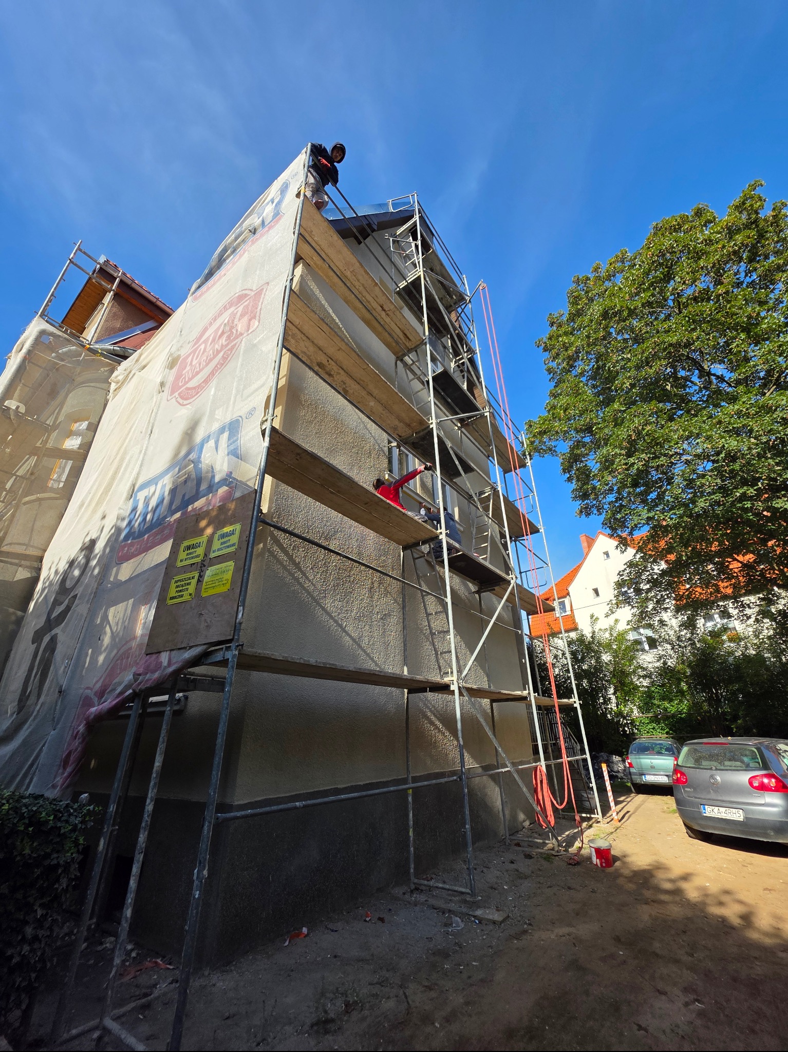 Ocieplanie budynku w trakcie prac. Rusztowanie z siatką ochronną, dwóch pracowników na różnych poziomach. Widok z dołu na elewację i fragment zieleni.