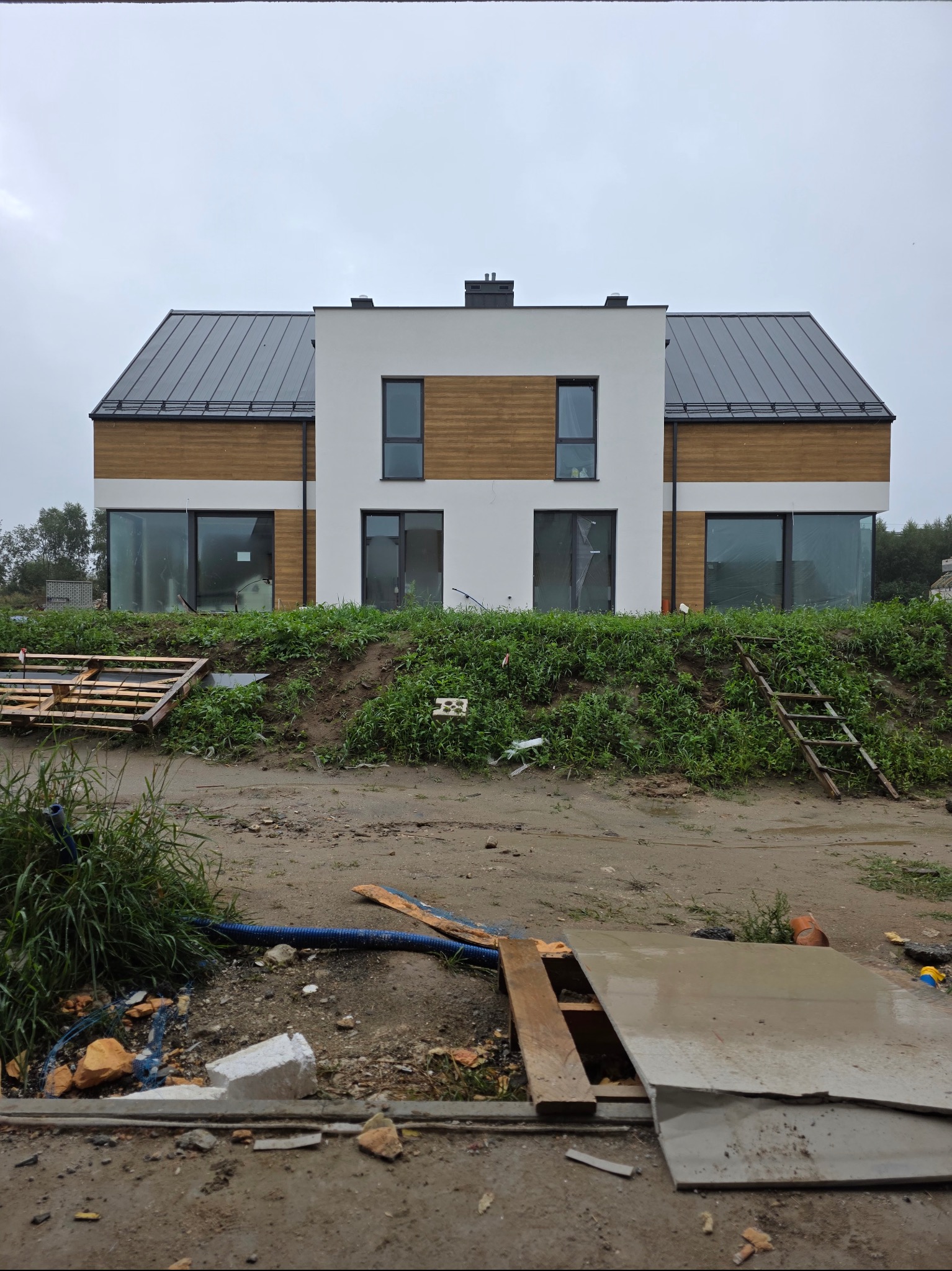 Nowoczesny dom z drewnianą elewacją i dużymi oknami, widoczny z zewnątrz w trakcie prac wykończeniowych. Na pierwszym planie gruz i materiały budowlane.