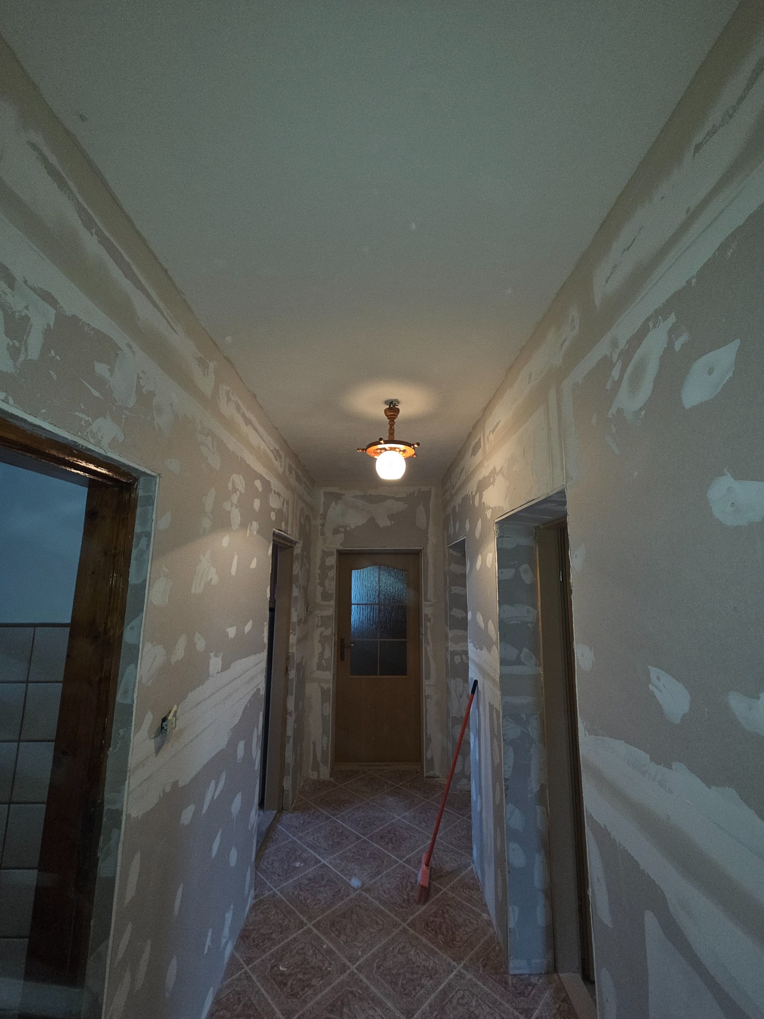 Korytarz w trakcie szpachlowania ścian, widoczne ślady gładzi, podłoga wyłożona płytkami w brązową kratę, w tle drzwi wejściowe i lampa sufitowa.
