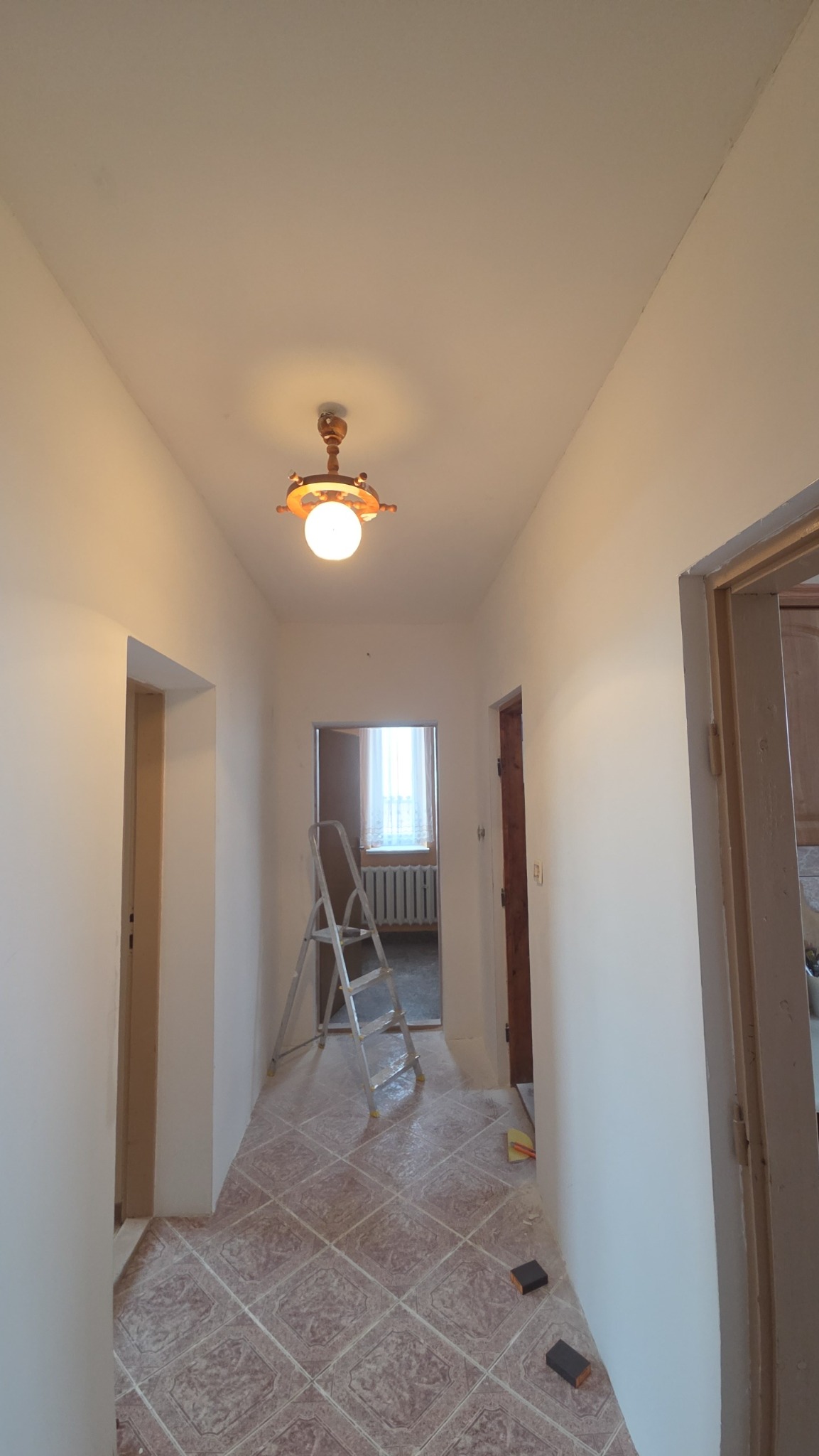 Korytarz po odświeżeniu ścian, widoczna drabina malarska, charakterystyczna lampa sufitowa i płytki podłogowe w romby. Świeżo pomalowane ściany na biało.