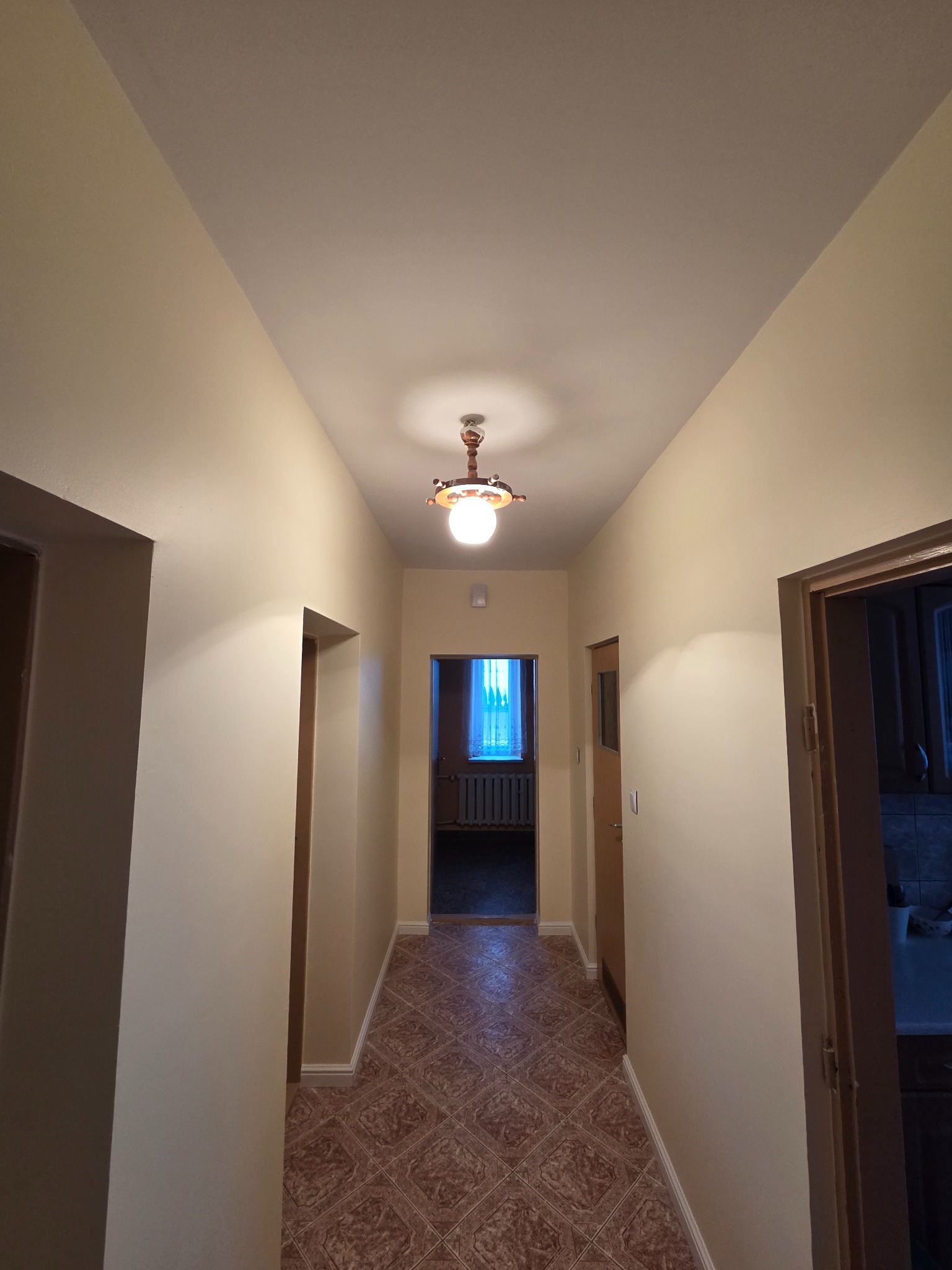 Jasny korytarz po remoncie z beżowymi ścianami, wzorzystą podłogą i oświetleniem sufitowym. Widoczne drzwi i fragment okna w tle. Neutralna kolorystyka.