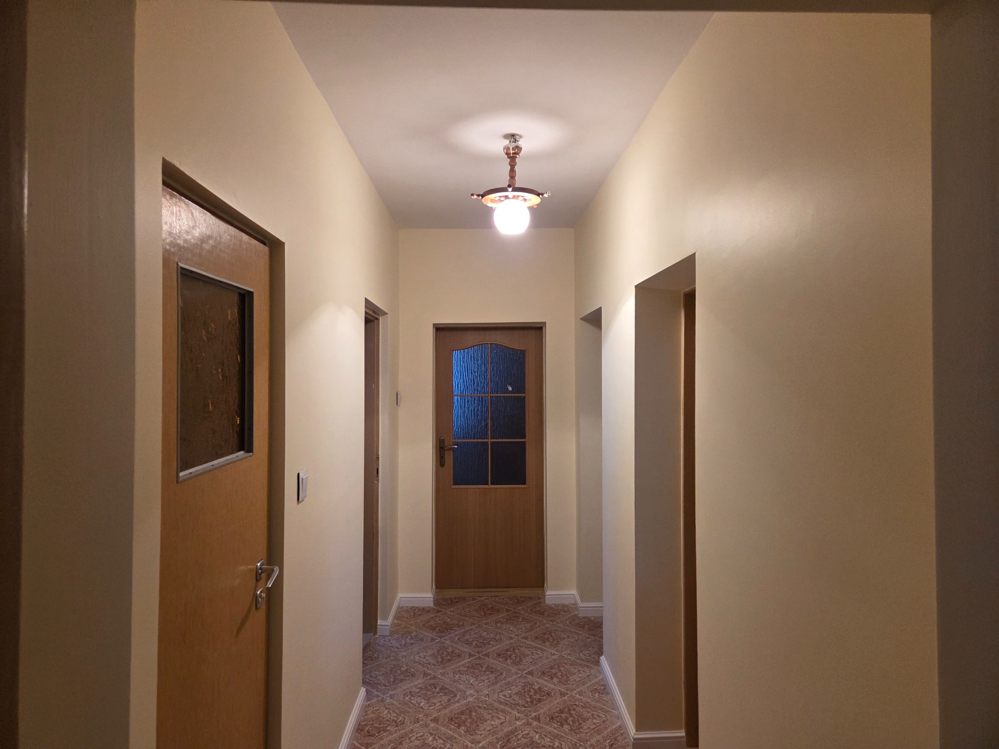 Korytarz po odświeżeniu: kremowe ściany, brązowe drzwi z szybami i wzorzysta podłoga. Centralnie umieszczona lampa sufitowa oświetla przestrzeń.