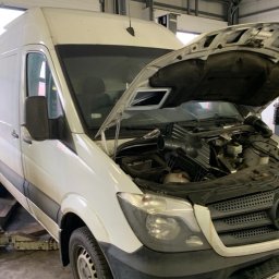 SILESIAMOTO SPÓŁKA Z OGRANICZONĄ ODPOWIEDZIALNOŚCIĄ - Biały van Mercedes na podnośniku w warsztacie, z otwartą maską ukazującą silnik. Widoczne elementy układu dolotowego i chłodzenia. Oświetlenie warsztatowe.