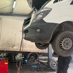 SILESIAMOTO SPÓŁKA Z OGRANICZONĄ ODPOWIEDZIALNOŚCIĄ - Podniesiony biały samochód dostawczy na podnośniku w warsztacie. Widoczne koła, opony, elementy zawieszenia i pracownik w trakcie wymiany opon. Warsztat z wyposażeniem.