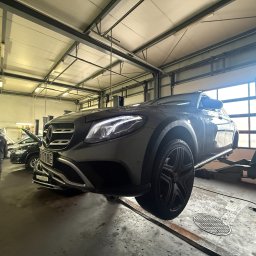 SILESIAMOTO SPÓŁKA Z OGRANICZONĄ ODPOWIEDZIALNOŚCIĄ - Szary Mercedes na podnośniku w warsztacie. Widoczne oświetlenie LED, inne auta w tle i pracownik. Ujęcie z dołu podkreśla wysokość podnoszenia.