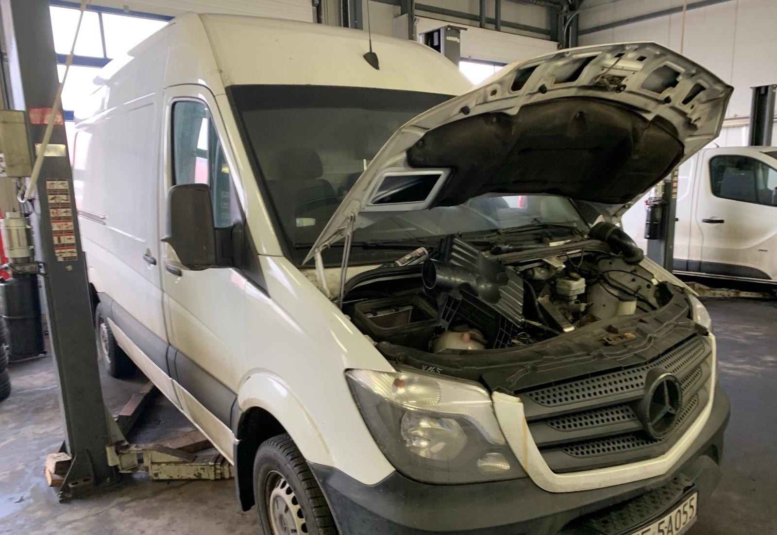 Biały van Mercedes na podnośniku w warsztacie, z otwartą maską ukazującą silnik. Widoczne elementy układu dolotowego i chłodzenia. Oświetlenie warsztatowe.