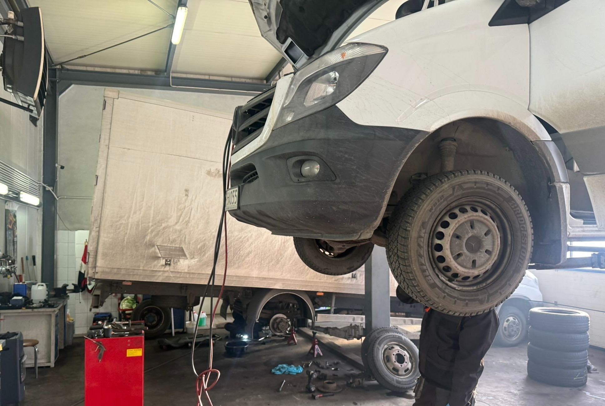 Podniesiony biały samochód dostawczy na podnośniku w warsztacie. Widoczne koła, opony, elementy zawieszenia i pracownik w trakcie wymiany opon. Warsztat z wyposażeniem.