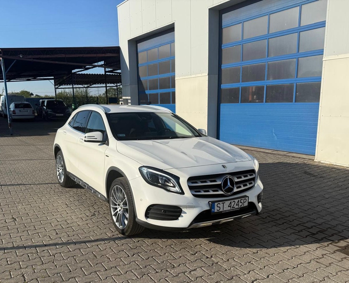 Biały Mercedes GLA na kostce brukowej przed warsztatem z niebieskimi bramami garażowymi w słoneczny dzień. Widoczny numer rejestracyjny.