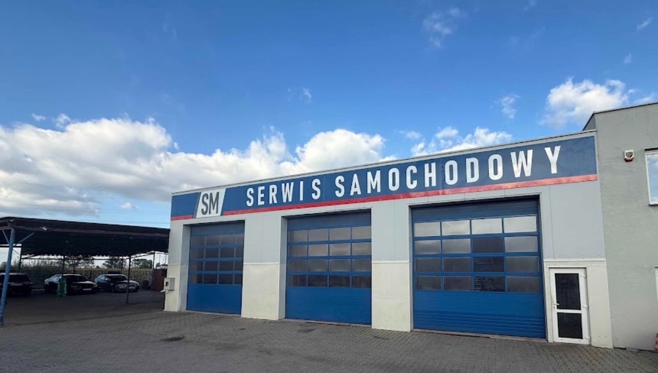 Fasada budynku serwisu samochodowego z trzema bramami garażowymi i napisem 'Serwis Samochodowy' na tle błękitnego nieba z chmurami.
