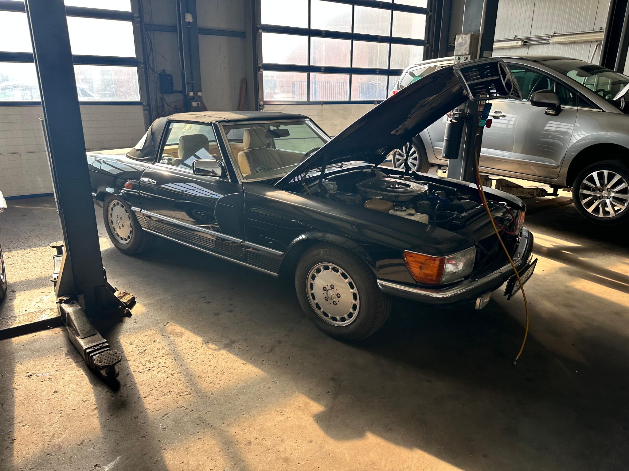 Czarny kabriolet z otwartą maską w warsztacie, podnośnik obok. Widoczne okna i inne pojazdy w tle. Diagnostyka silnika w trakcie naprawy.