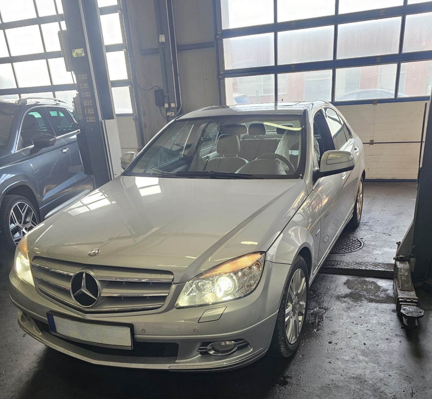 Srebrny Mercedes-Benz C-Klasa na podnośniku w warsztacie, z widocznymi detalami karoserii i oświetlenia. Duże okna warsztatu w tle.