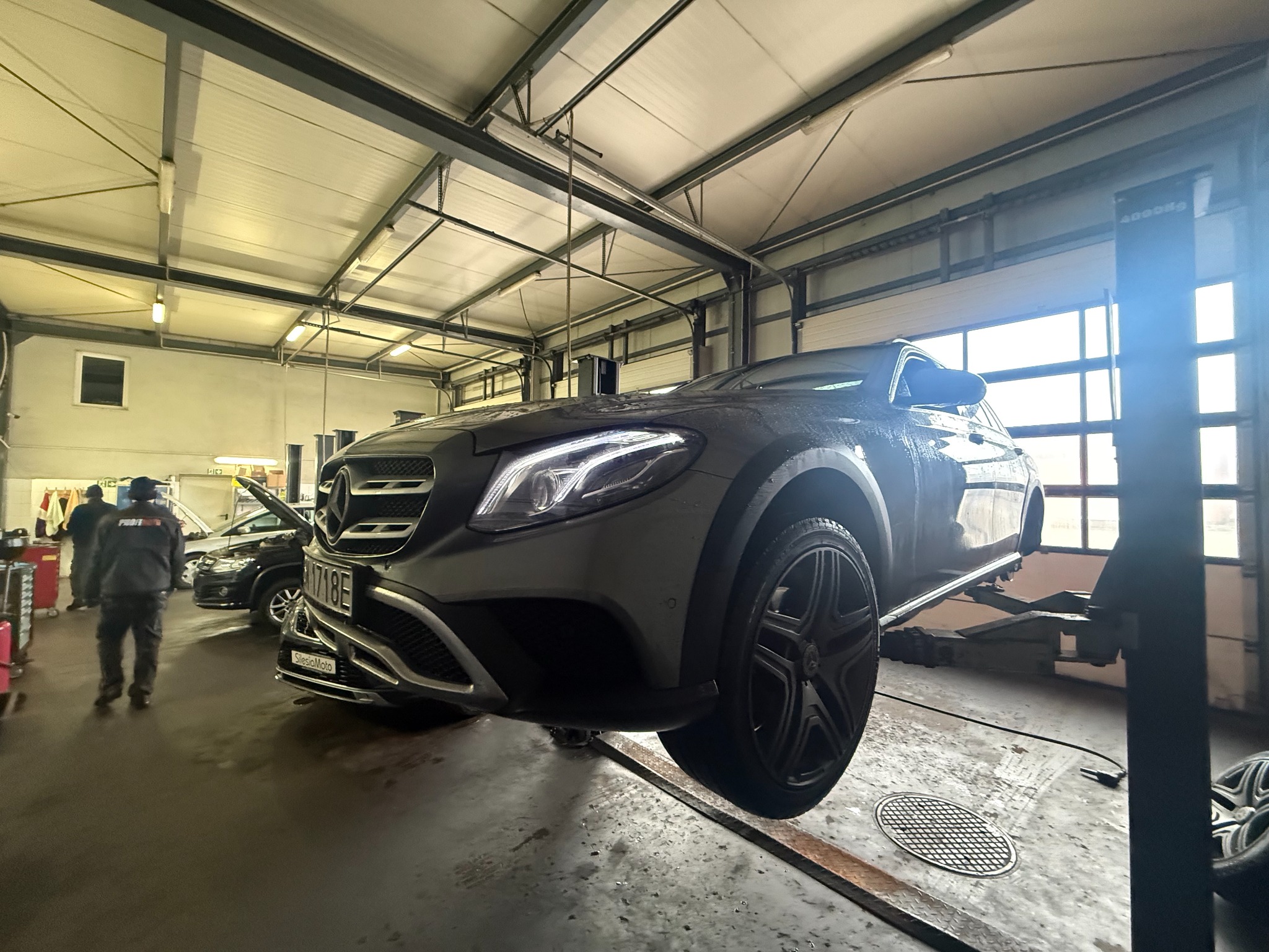 Szary Mercedes na podnośniku w warsztacie. Widoczne oświetlenie LED, inne auta w tle i pracownik. Ujęcie z dołu podkreśla wysokość podnoszenia.