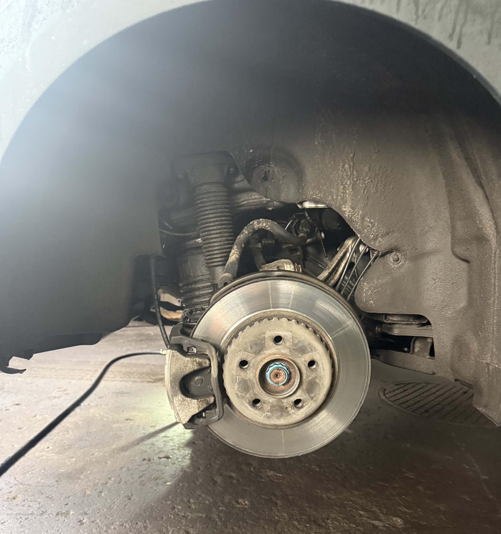 Tarcza hamulcowa z zaciskiem w samochodzie na podnośniku, widoczne elementy zawieszenia i przewody. Ujęcie z bliska, oświetlenie warsztatowe.