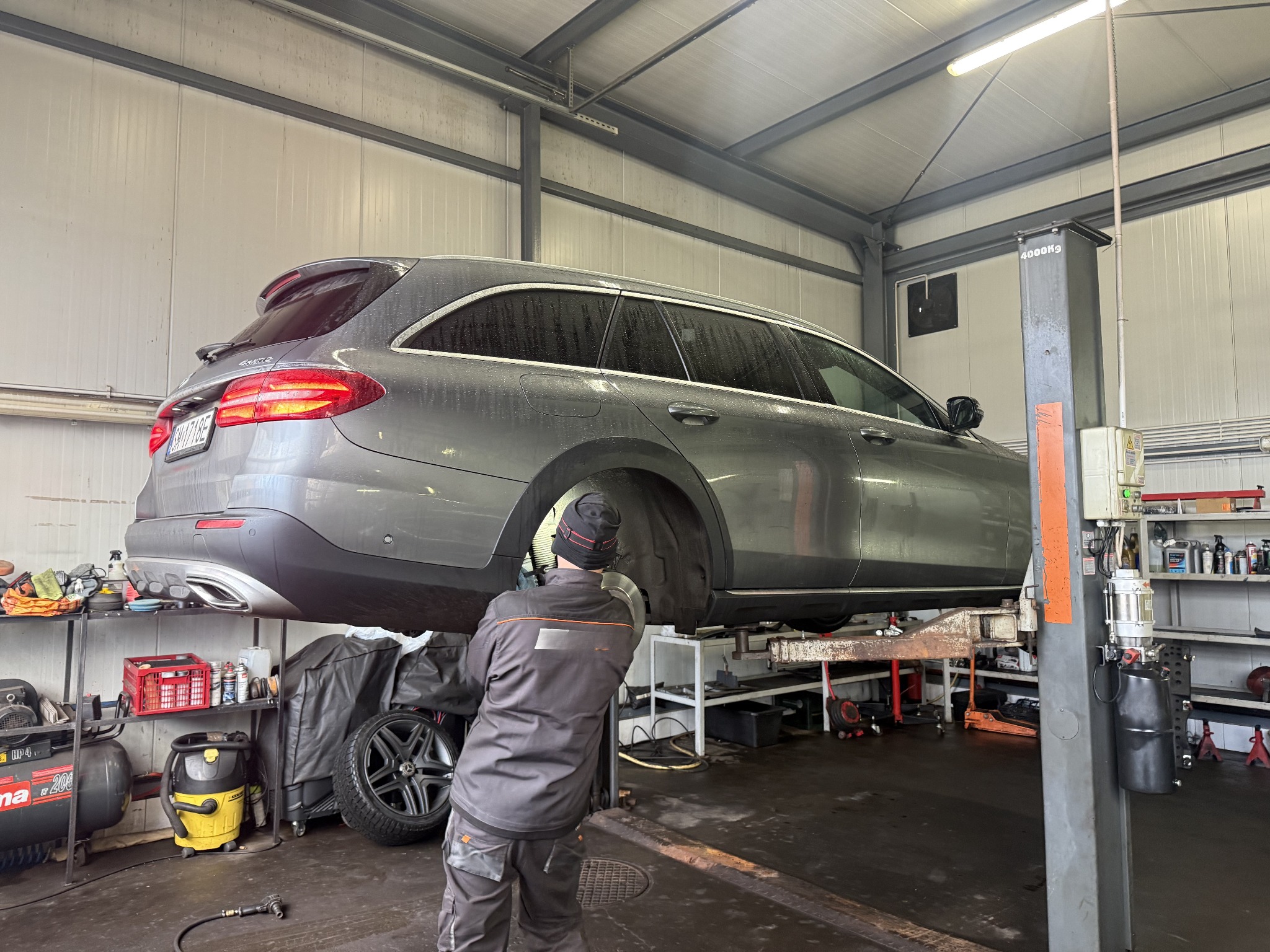 Szary samochód kombi na podnośniku w warsztacie. Widoczny mechanik w kombinezonie przy kole, półki z narzędziami i akcesoriami. Oświetlenie warsztatowe.