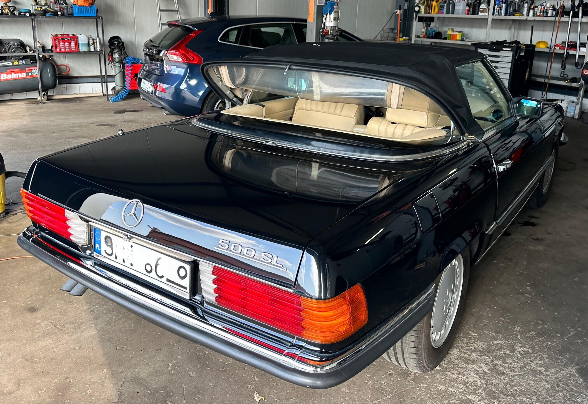 Czarny Mercedes 500 SL kabriolet z beżowym wnętrzem, zaparkowany w warsztacie. Widoczny fragment innego auta i regał z narzędziami w tle. Klasyczny model.