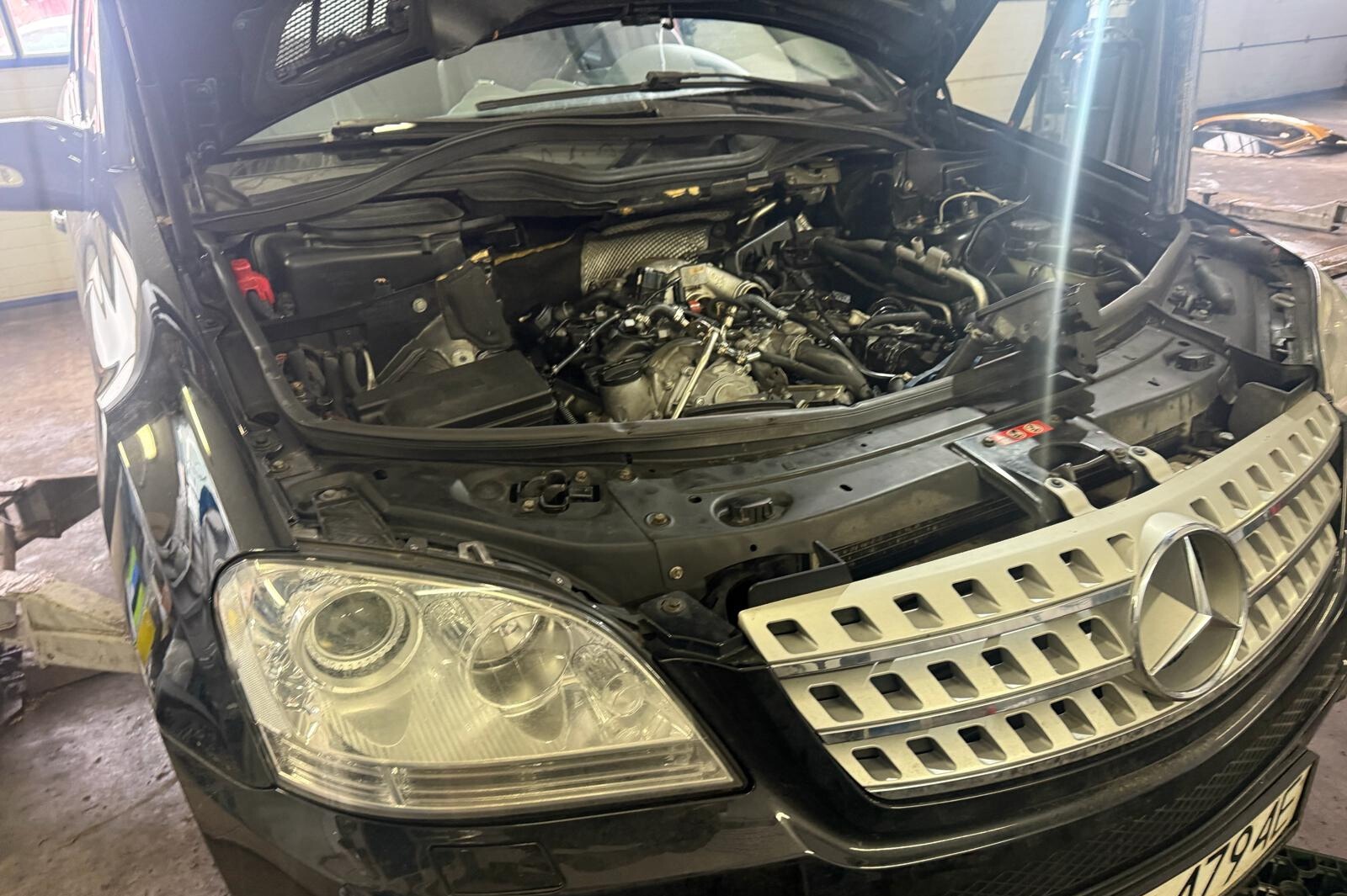 Czarny samochód z otwartą maską, widoczny silnik w trakcie naprawy. Detal: srebrna atrapa chłodnicy z gwiazdą Mercedesa. Warsztat samochodowy w tle.