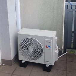 Beskidklima Mateusz Wawrzyk - Biała jednostka zewnętrzna klimatyzacji Sinclair DC Inverter na czarnych podkładkach, ustawiona na szarym bruku przy ścianie budynku.