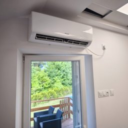 Beskidklima Mateusz Wawrzyk - Biały klimatyzator Sinclair zamontowany nad oknem balkonowym z widokiem na zieleń. Widoczne gniazdka elektryczne i fragment sufitu podwieszanego.