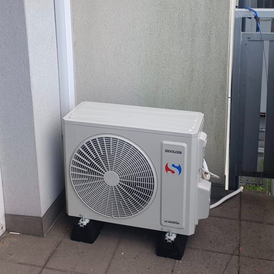 Biała jednostka zewnętrzna klimatyzacji Sinclair DC Inverter na czarnych podkładkach, ustawiona na szarym bruku przy ścianie budynku.