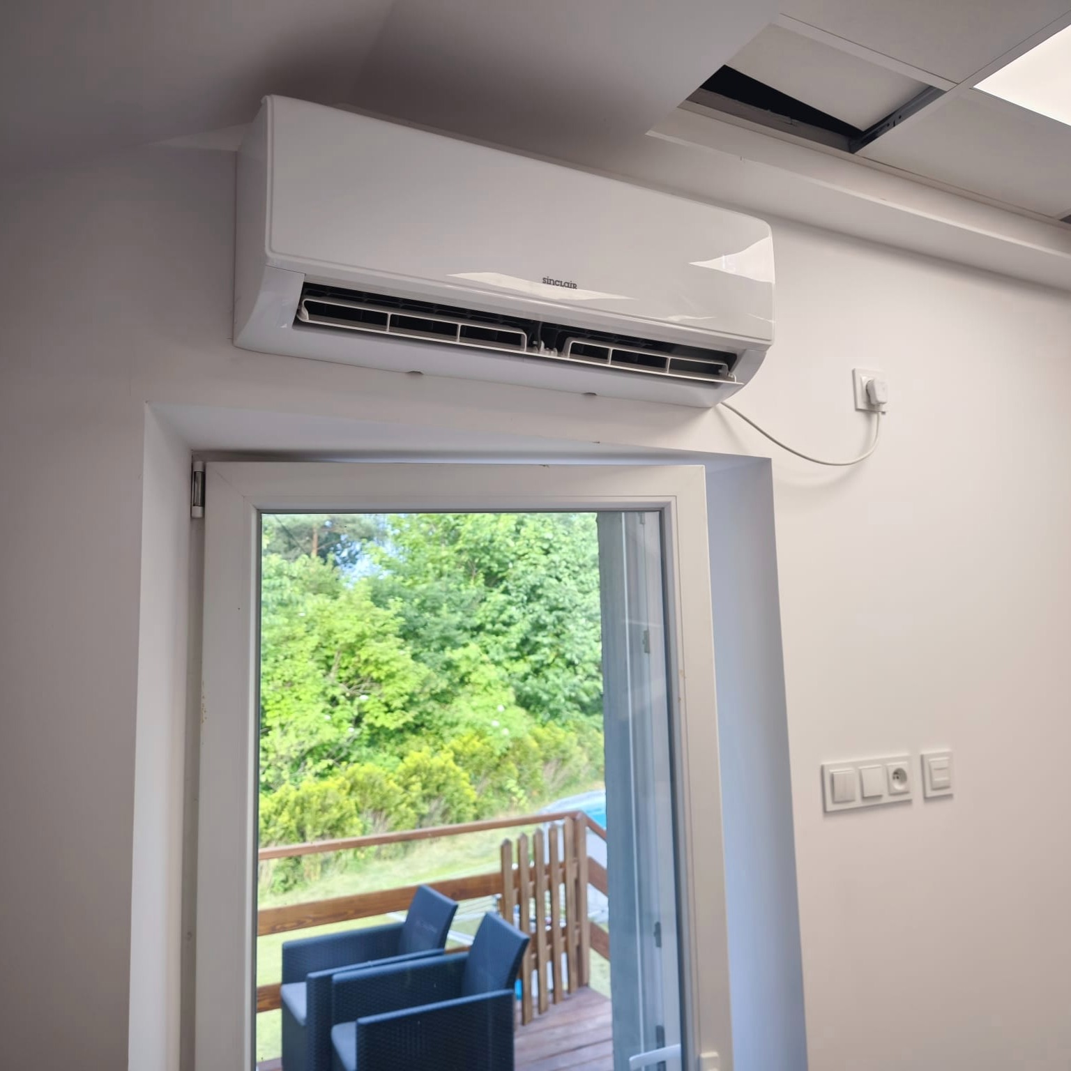 Biały klimatyzator Sinclair zamontowany nad oknem balkonowym z widokiem na zieleń. Widoczne gniazdka elektryczne i fragment sufitu podwieszanego.