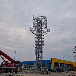 SZYMSTAL SZYMON SĄDEJ - Wysoka, stalowa konstrukcja w trakcie montażu, widoczny dźwig i ekipa budowlana na tle budynku z niebiesko-żółtą elewacją, pochmurne niebo.