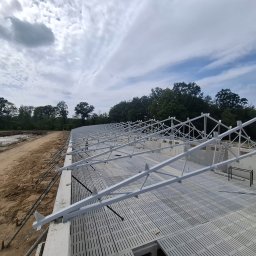 SZYMSTAL SZYMON SĄDEJ - Konstrukcja hali w trakcie budowy: białe, metalowe elementy szkieletu na betonowym fundamencie, widok z góry, w tle drzewa i pochmurne niebo.