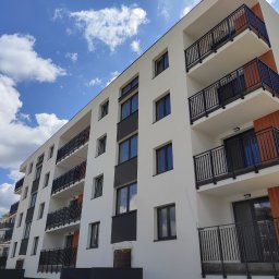 SZYMSTAL SZYMON SĄDEJ - Nowoczesny budynek wielorodzinny z balkonami zabezpieczonymi czarnymi, metalowymi balustradami na tle błękitnego nieba z chmurami. Ujęcie z dołu, podkreślające wysokość budynku.