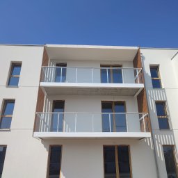 SZYMSTAL SZYMON SĄDEJ - Nowoczesny budynek z białymi balkonami i szklanymi balustradami, drewniane elementy dekoracyjne, minimalistyczny design, błękitne niebo w tle.