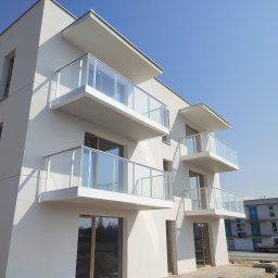 SZYMSTAL SZYMON SĄDEJ - Nowoczesny budynek z balkonami z balustradami szklanymi, widok z dołu, jasne tło nieba. W tle budynek w trakcie budowy i samochód dostawczy.