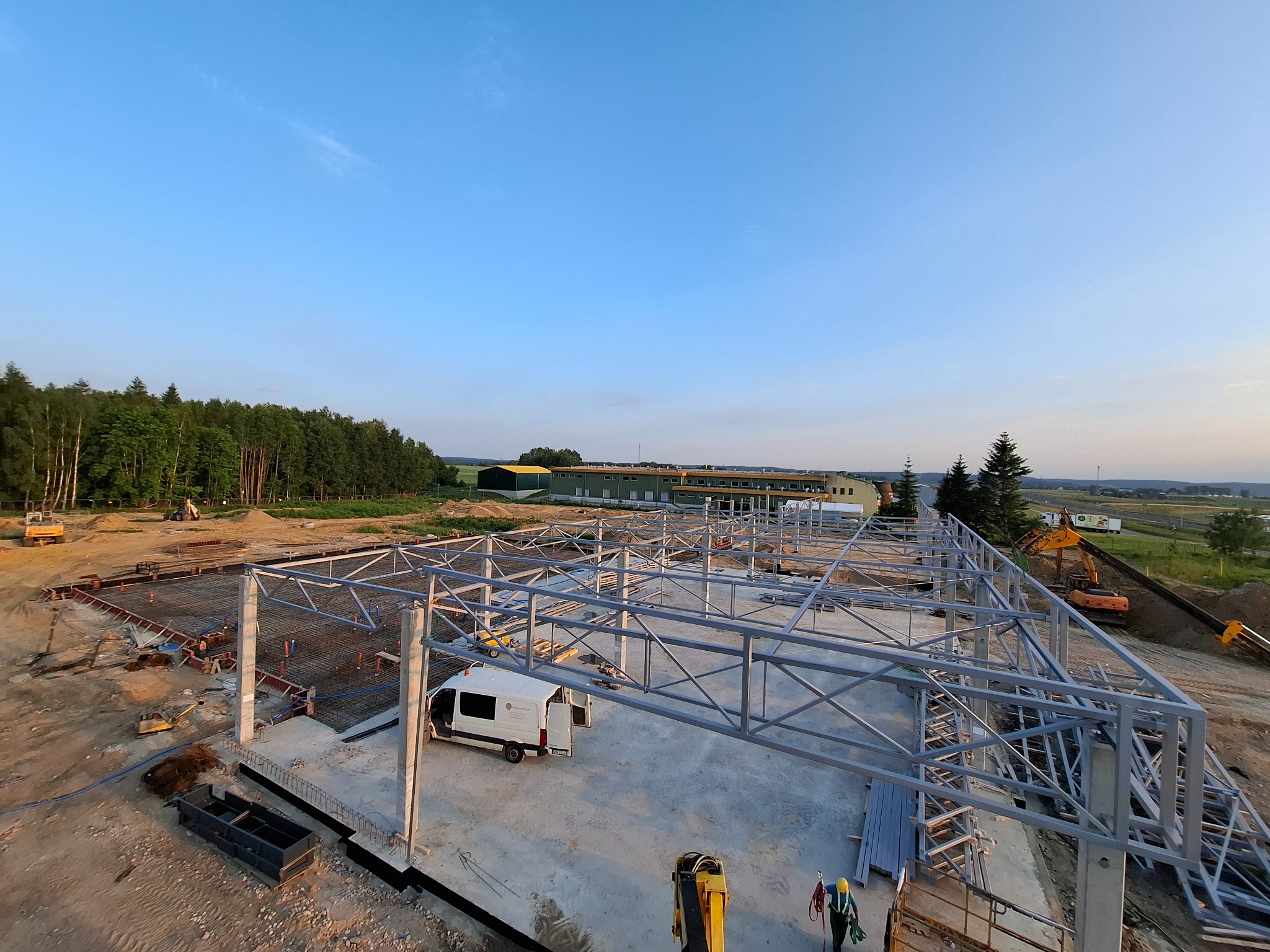Szkielet hali w budowie, widok z góry. Stalowa konstrukcja na betonowym fundamencie, w tle las i błękitne niebo. Na placu budowy stoi biały bus i pracuje koparka.