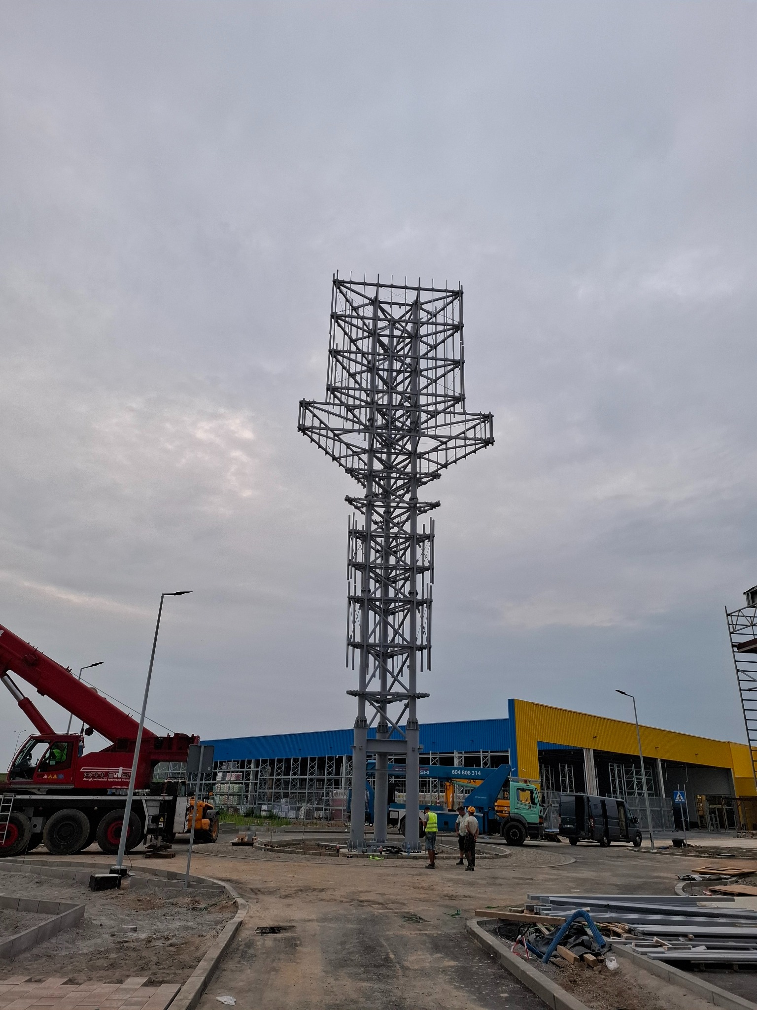 Wysoka, stalowa konstrukcja w trakcie montażu, widoczny dźwig i ekipa budowlana na tle budynku z niebiesko-żółtą elewacją, pochmurne niebo.