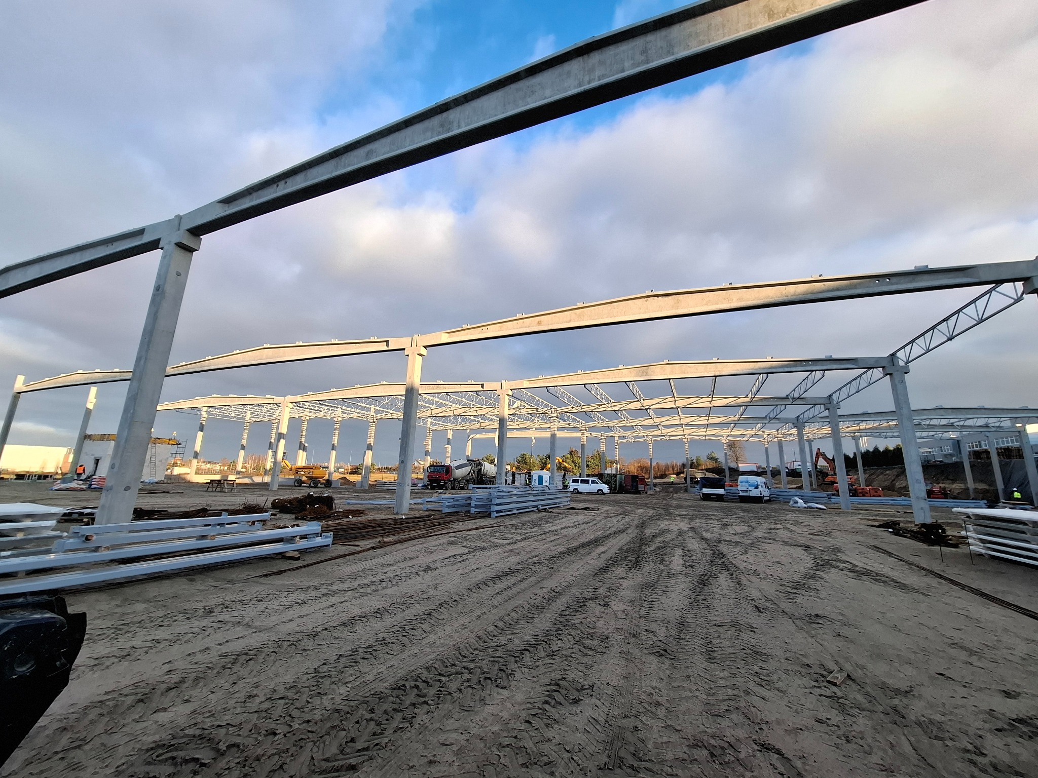 Wznoszenie konstrukcji stalowej hali: widok z dołu na szkielet budynku z betonowych słupów i metalowych belek, na tle pochmurnego nieba. W tle pojazdy budowlane.