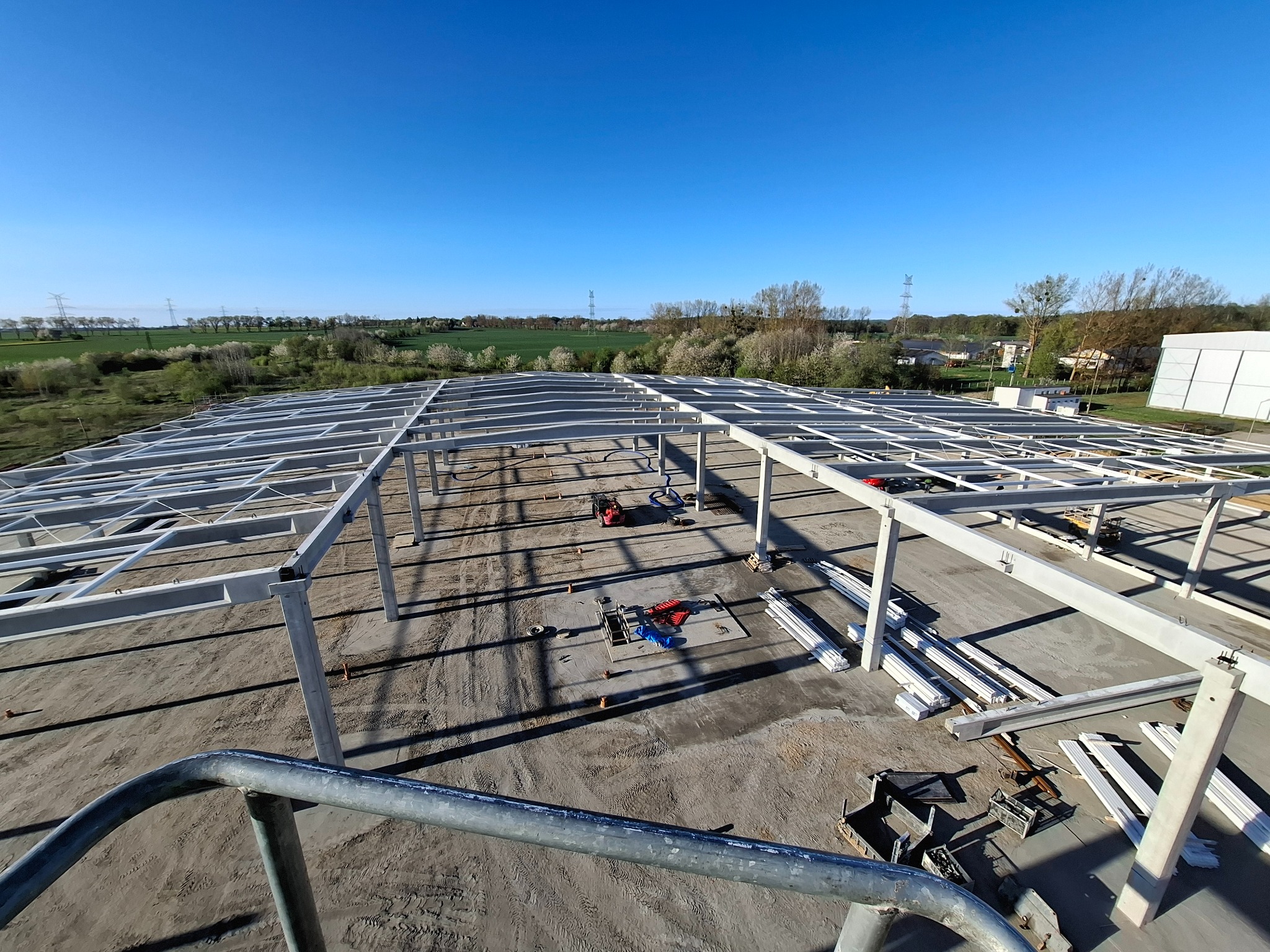 Wysoka perspektywa na szkielet hali w budowie, z widocznymi elementami konstrukcyjnymi i sprzętem budowlanym na tle zieleni i błękitnego nieba.