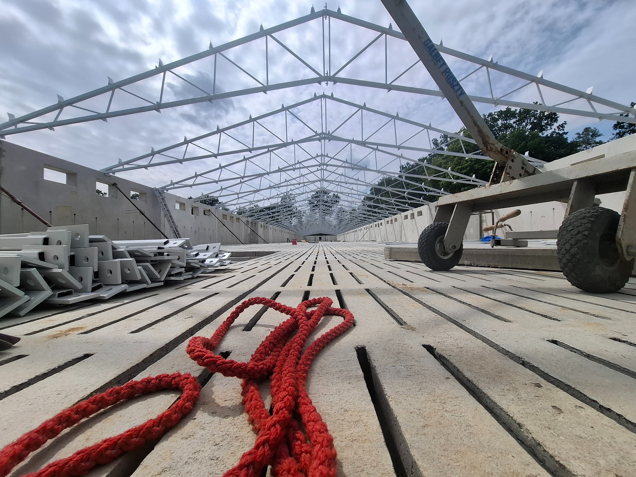 Wnętrze hali w budowie z widoczną konstrukcją stalową dachu, betonowymi ścianami z otworami okiennymi, elementami konstrukcyjnymi i czerwoną liną na podłożu.